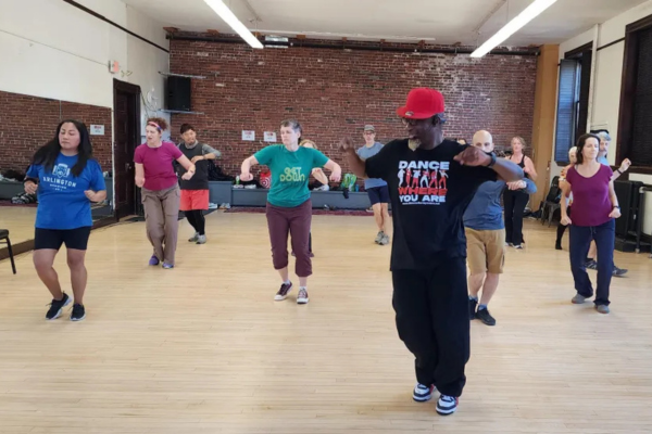 Man in black clothing and red hat dances in a dance studio with several students behind him